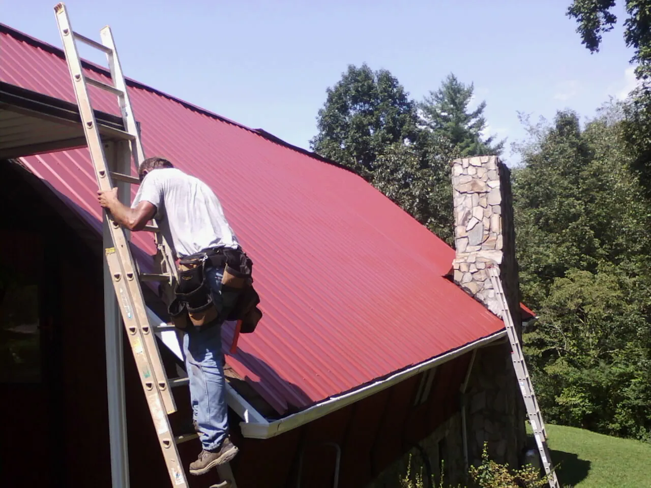 Roofer on a ladder beside a red metal roof and stone chimney — Soffit & Fascia Repair by Roofing Brothers in Mabank